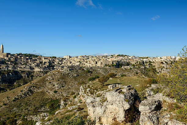 Expansive view of Matera, Italy, with its ancient stone buildings perched atop rugged cliffs and a sunlit rocky valley in the foreground beneath a clear sky.