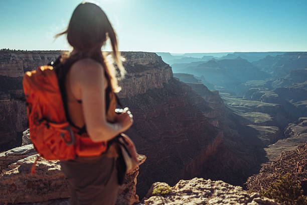 Female hiker with backpack standing on a viewpoint overlooking the Grand Canyon. Travel, adventure, hiking, and outdoor exploration concept in a natural desert landscape under a clear blue sky.