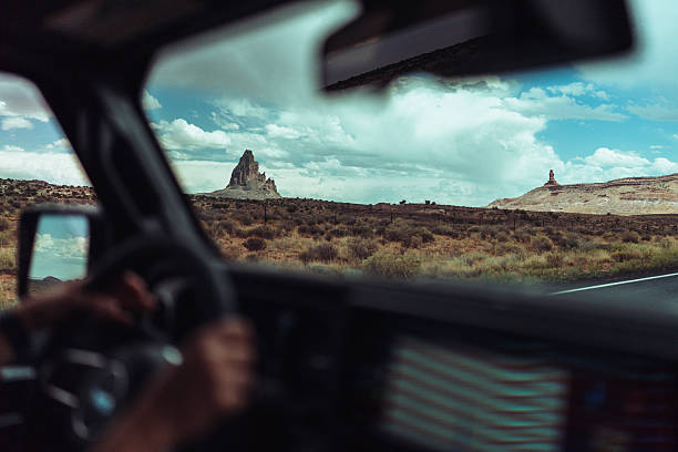 View from inside a moving vehicle looking out across an open desert plain with iconic sandstone buttes under a dramatic sky. The framing through the windshield conveys travel, motion, and the immersive experience of a road trip through the American Southwest.