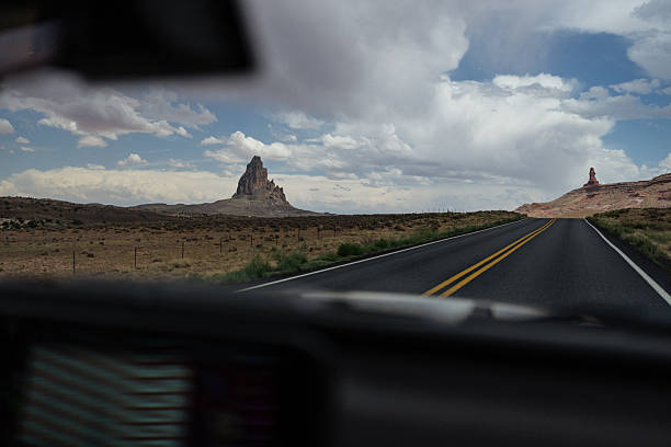 View from inside a moving vehicle looking out across an open desert plain with iconic sandstone buttes under a dramatic sky. The framing through the windshield conveys travel, motion, and the immersive experience of a road trip through the American Southwest.