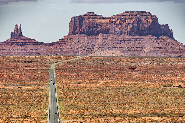 A straight highway stretches toward the iconic sandstone buttes of Monument Valley, cutting through a vast desert plain under a wide blue sky. The scene evokes freedom, distance, and the timeless scale of the American Southwest.