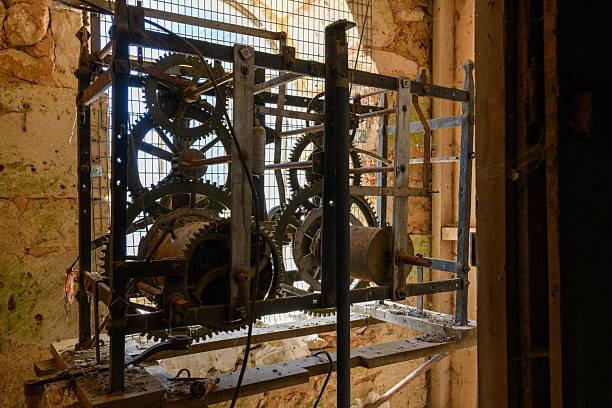 A vintage iron clockwork mechanism with interlocking gears stands inside a weathered stone tower, illuminated by soft daylight filtering through a barred window. The scene highlights intricate engineering, aged metal, and the textured masonry of historic architecture.