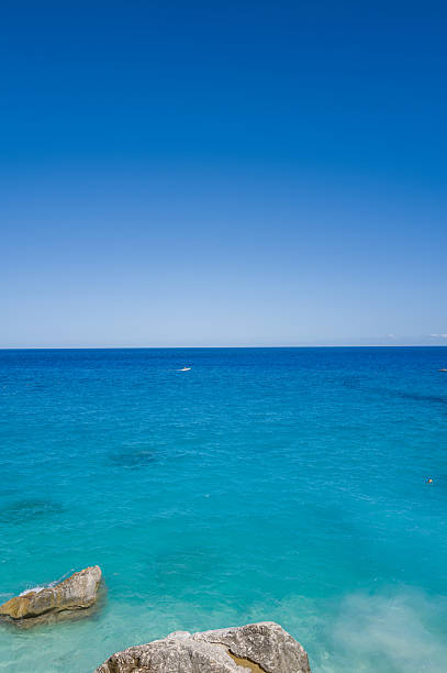 Vivid turquoise water laps against sunlit coastal rocks beneath a cloudless deep blue sky at Cala Goloritze, Sardinia. The tranquil seascape radiates clarity and calm, perfect for summer relaxation.