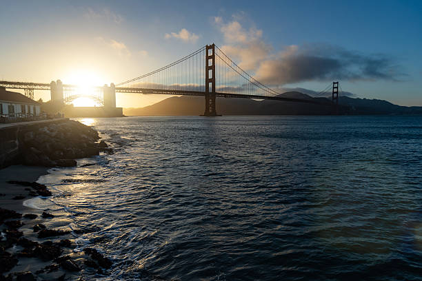 Wide view of the Golden Gate Bridge at sunset, with warm evening light reflecting on the water of San Francisco Bay, rocky shoreline in the foreground, and low clouds over the surrounding hills.