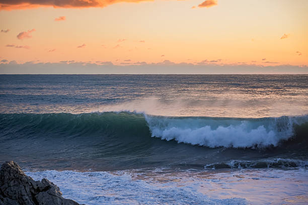 Wave fully curling and breaking over the sea at sunset. Orange evening sky, dramatic motion, and cold winter water create a dynamic coastal scene.