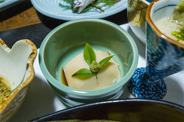 A smooth block of tofu is served in a pale green ceramic bowl, topped with a fresh leaf and wasabi. The arrangement features gentle lighting and a refined, minimalist presentation among other Japanese dishes.