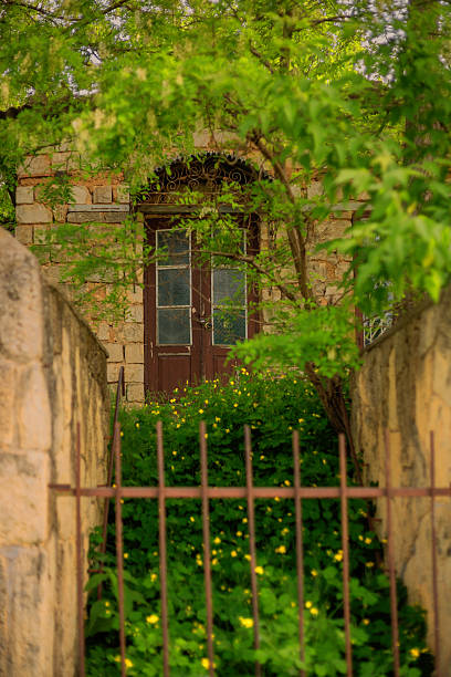 Traditional stone building featuring an arched doorway, rusty iron gate, and vibrant green foliage