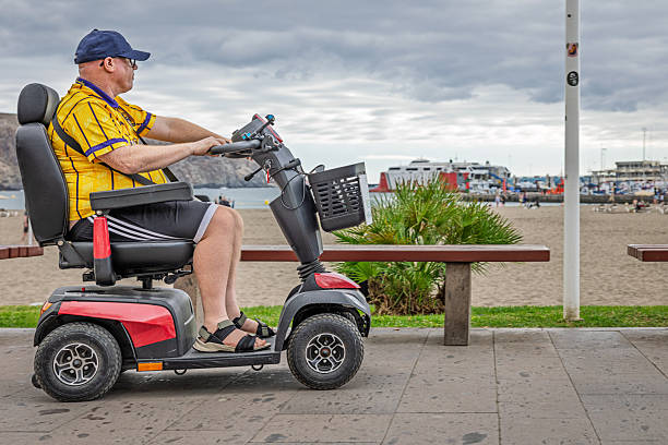 Los Christianos, Tenerife, Spain, Europe - December 19th 2025: An elderly man uses a mobility scooter along the south coast of Tenerife in the Canary Islands. The area is part of one of the island’s long‑established tourist zones, supported by a professional tourism industry that for many years has provided mobility aids and accessibility services for visitors with disabilities.