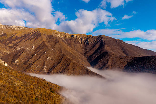 Wide view of a rugged mountain ridge covered in autumn vegetation, rising above a layer of low clouds under a blue sky with scattered clouds.