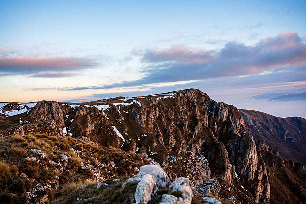 Rugged mountain ridge with rocky formations and patches of snow, captured under a calm pastel-colored evening sky with distant clouds.