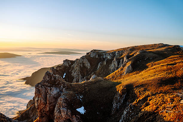 Rocky mountain ridge glowing in golden-hour light, overlooking distant cloud-covered valleys and layered hills.