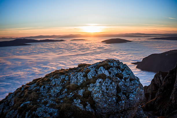 Foreground rocky outcrop framing a wide view of a valley completely filled with clouds under soft sunrise tones.