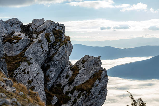 Sharp rocky formations overlooking layered mountain valleys filled with low clouds under soft daylight.