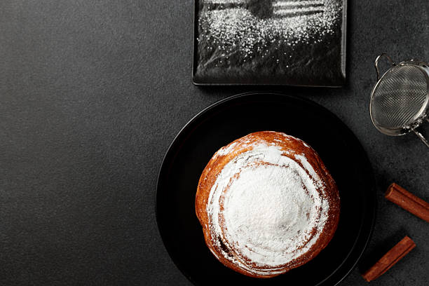 Round, delicious bun with cinnamon and powdered sugar on a black plate on a gray background