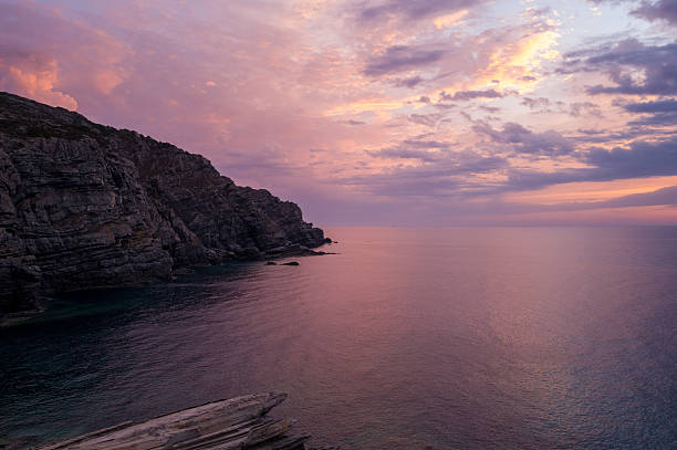 Warm pink and violet hues illuminate dramatic stone cliffs overlooking calm Mediterranean waters at dusk near Stintino, Sardinia. The tranquil scene features textured rock faces and a softly colored sky.