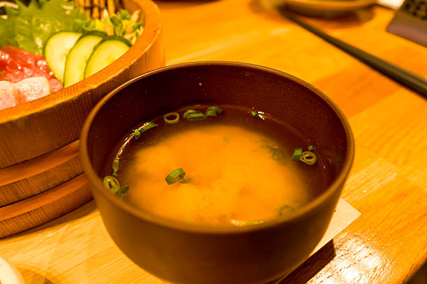 A steaming bowl of miso soup garnished with sliced green onions sits on a wooden table, next to a traditional wooden tub of sashimi. Warm lighting highlights the comforting, rustic atmosphere.
