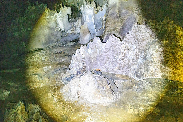 Sharp-edged limestone formations and textured mineral deposits are illuminated by a flashlight inside the shadowy interior of Phu Kham Cave, Laos. The rugged surfaces and layered rock patterns highlight the dramatic geology of this karst environment.