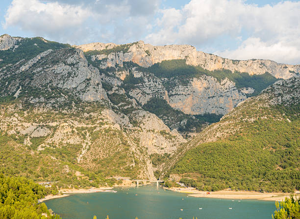 Vivid turquoise water meets sandy shores beneath dramatic limestone peaks and lush green hills in the Gorges du Verdon, with sunlight casting soft shadows across the landscape.
