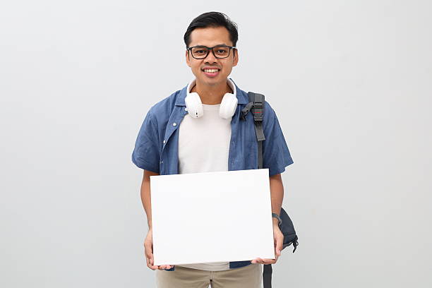 istock Young Asian male student wearing backpack, headphone around neck, holding blank board, standing smiling looking at camera, isolated white background. 2255885500