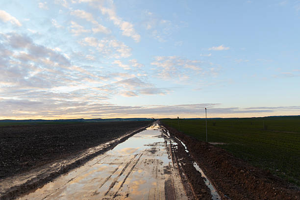 Muddy dirt road with puddles reflecting the sky after rain in rural farmland. Atmospheric countryside scene with clouds, water reflections and natural textures.