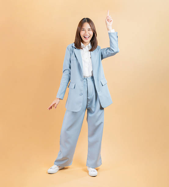 Cheerful young woman in a blue suit expressing excitement as points upward, standing confidently against a beige background.