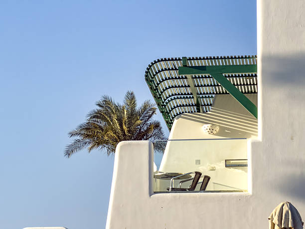 Modern white building with balcony and palm tree against clear blue sky. Minimalist architectural detail in a sunny coastal environment.