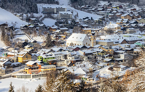 A breathtaking aerial view of Werfen, Austria, blanketed in a thick layer of snow during winter. The charming village showcases traditional Austrian architecture with colorful houses and a prominent church with a striking yellow steeple. The surrounding landscape is a pristine white, creating a picturesque and serene winter scene. The image captures the beauty and tranquility of a snow-covered alpine village.