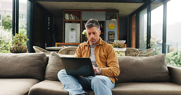 istock Remote work, laptop and mature man on sofa for online article, website research and proofreading. Freelancer, living room and person on computer for editing report, story review and planning in home 2255893568