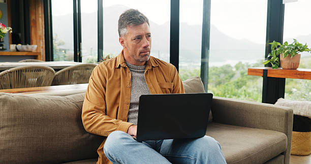 istock Thinking, laptop and mature man on sofa for online article, website research and proofreading. Freelancer, living room and person on computer for editing report, story review and remote work in home 2255893613