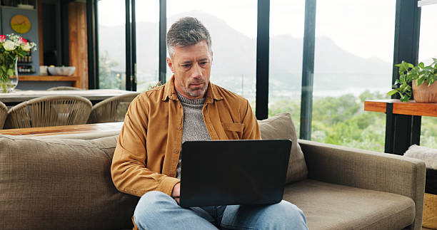 istock Remote work, laptop and mature man in home for online article, website research and proofreading. Freelancer, living room and person on computer for editing report, story review and planning on sofa 2255893633