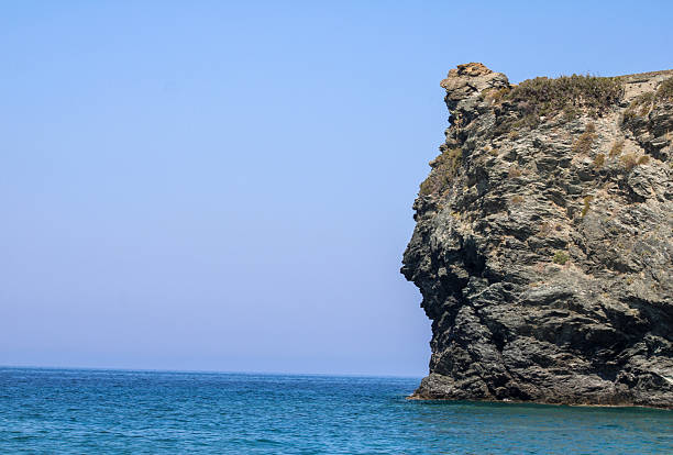 a dramatic coastal scene featuring a rugged rocky cliff jutting out into the calm blue sea, with sparse vegetation growing on top, under a clear sky with soft gradient tones, creating a natural and minimalistic seascape