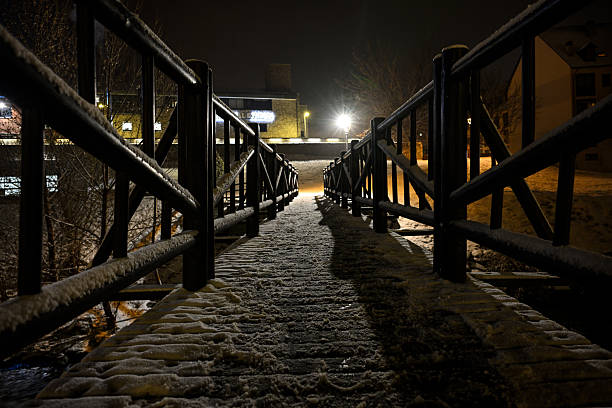 The image depicts a snow-covered pedestrian bridge at night, framed by wooden railings and illuminated from behind by ambient street and building lights. The interplay of shadow and glow creates a dramatic contrast, highlighting the bridges symmetry and the textured snow. The scene evokes solitude, warmth, and quiet movement through a winter urban landscape.