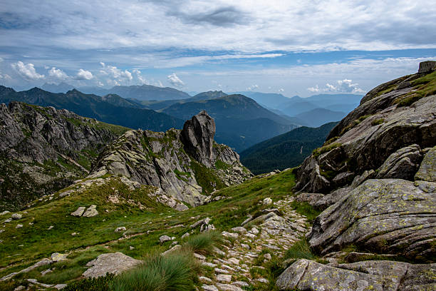 Rocky alpine ridge with a lone vertical peak rising above green slopes and distant mountain layers in the Lagorai range of Trentino, Italy, under a bright summer sky.
