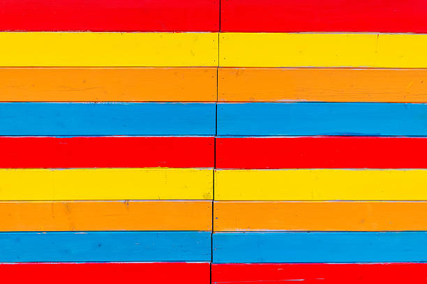 Colorful horizontal stripes in red, blue, yellow, and orange adorn a wooden wall on a bright afternoon, bringing cheer and creativity to the surroundings.