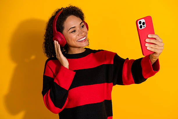 Young woman in striped pullover with red headphones taking a cheerful selfie against bright yellow background