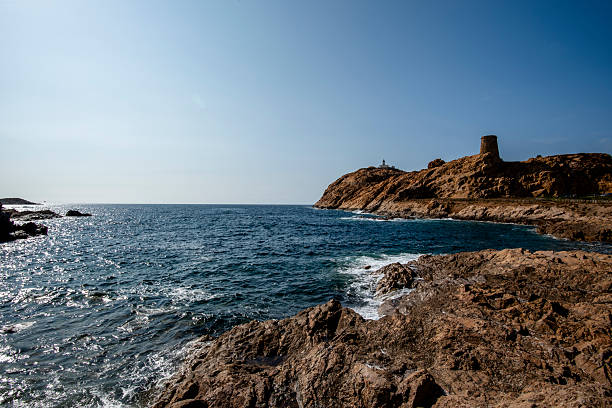 Rocky Corsican shoreline with deep blue waves, a coastal tower and a distant lighthouse perched on golden cliffs under a bright summer sky.