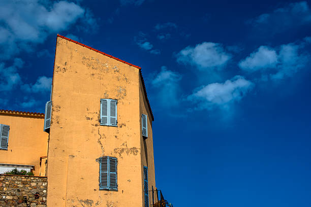 Yellow Mediterranean house facade with blue shutters against deep blue sky and clouds in Corsica, France. Minimal architecture, rustic texture and calm southern European atmosphere.