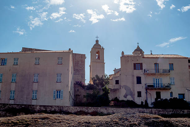 Historic village buildings with church bell tower under bright sky in Corsica, France. Mediterranean architecture, pastel facades, religious landmark and serene southern European atmosphere.