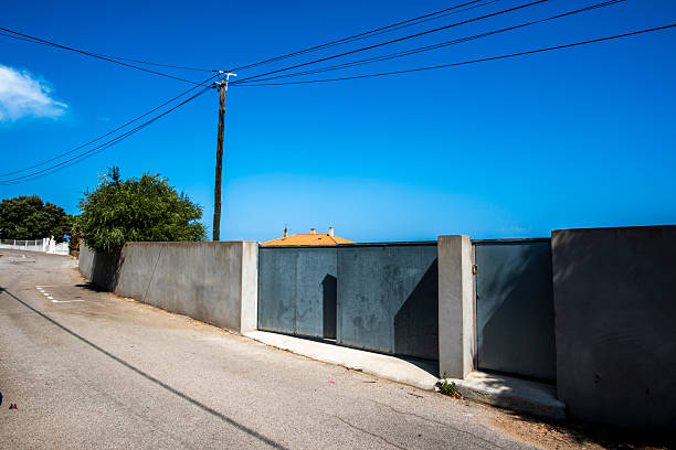 Quiet residential street with concrete wall, metal gate and power lines under clear blue sky in Corsica, France. Minimal suburban landscape, strong lines and calm Mediterranean atmosphere.