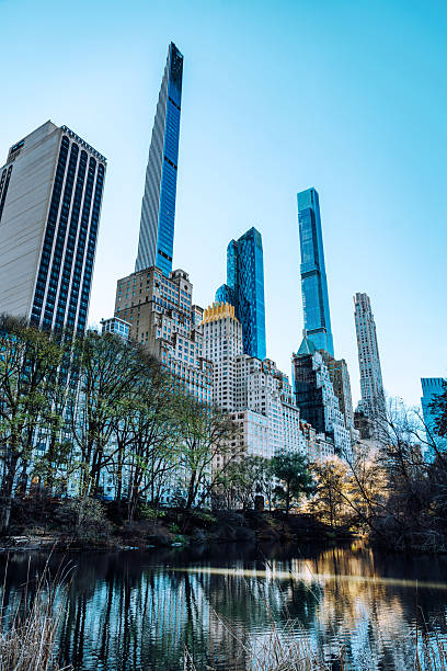 A dramatic cityscape of Manhattan's iconic skyscrapers rising above a tranquil park and reflective water. Bright blue sky and glass façades frame the urban waterfront scene with trees along the shore.