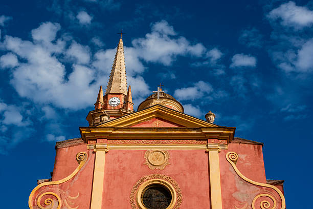 Colorful church facade with clock tower and spire against dramatic blue sky in Corsica, France. Baroque Mediterranean architecture, symmetry, heritage and southern European atmosphere.