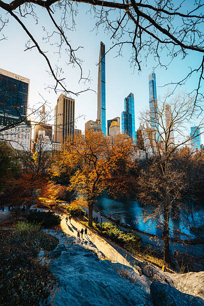 Sunlit autumn park scenes beside the river frame a historic Manhattan skyline. A peaceful urban escape with golden trees, calm water, and people strolling along a shaded path.