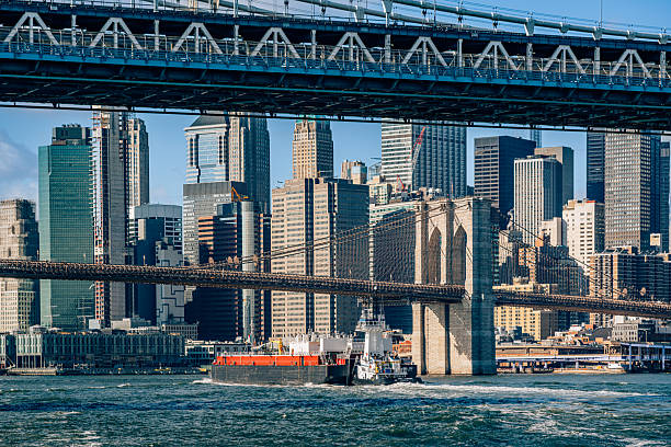 A sweeping view of the Manhattan skyline framed by a large suspension bridge overhead. A cargo ship glides through the harbor as towering skyscrapers rise in the New York City skyline.