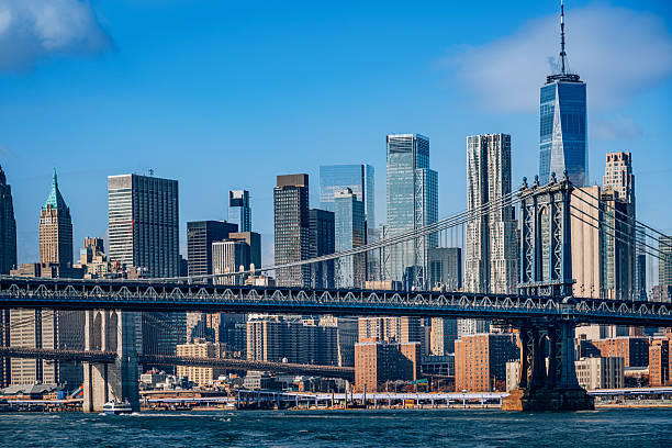 A sweeping view of Manhattan's skyline with the Brooklyn Bridge spanning the East River. Bright blue sky, glass towers, and urban energy convey iconic city life and architectural grandeur.