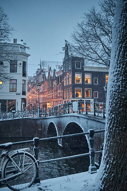 Amsterdam canals and typical canal houses in the old part of the city during a winter morning. The streets are covered in white from the snow, the city lights are on and the sky is grey because of the clouds. You can see some snow falling down.