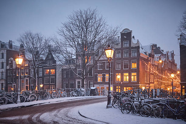 Amsterdam canals and typical canal houses in the old part of the city during a winter morning. The streets are covered in white from the snow, the city lights are on and the sky is grey because of the clouds. You can see some snow falling down.