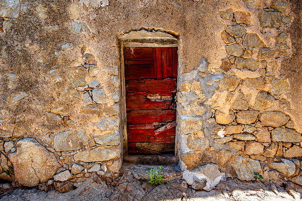 Weathered red wooden door set in a rough stone wall in Corsica, France. Peeling paint, cracked textures and natural light evoke abandonment, resilience and timeless Mediterranean history.