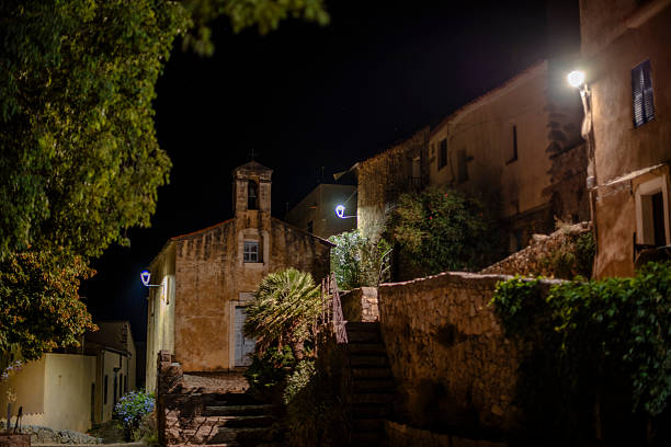 Night view of a historic Corsican village street with stone houses, church facade and stairs illuminated by street lamps. Quiet Mediterranean atmosphere, European heritage travel scene.