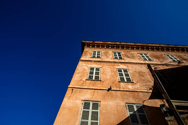Mediterranean residential building facade in Corsica, France, seen from below against deep blue sky. Warm plaster walls, shuttered windows and southern European architectural character.