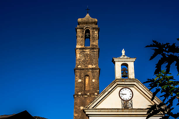 Historic church bell tower and clock facade in Corsica, France, under deep blue sky. Mediterranean stone architecture, religious heritage and timeless European village atmosphere.